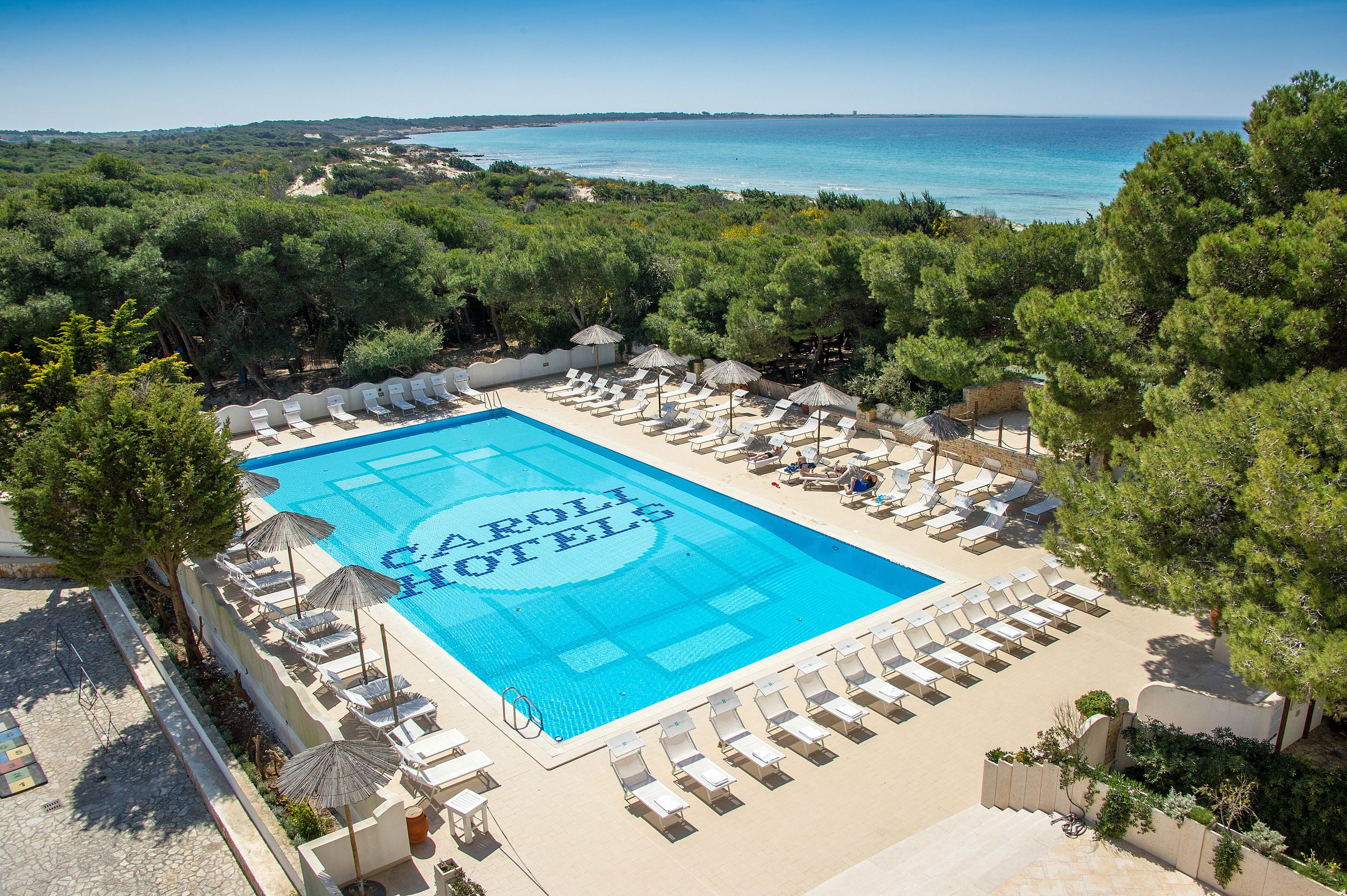 A pool area with lounge chairs and umbrellas, surrounded by trees, with a view of the sea.