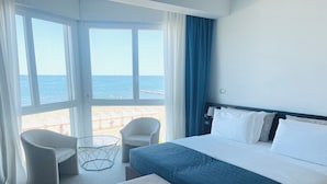 Habitación de hotel con cama, dos sillas, una mesita y vistas al mar a través de amplias ventanas.