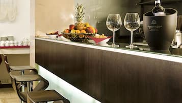 Un espace bar moderne avec un comptoir en verre, des tabourets de bar, un présentoir de fruits et une bouteille de vin.