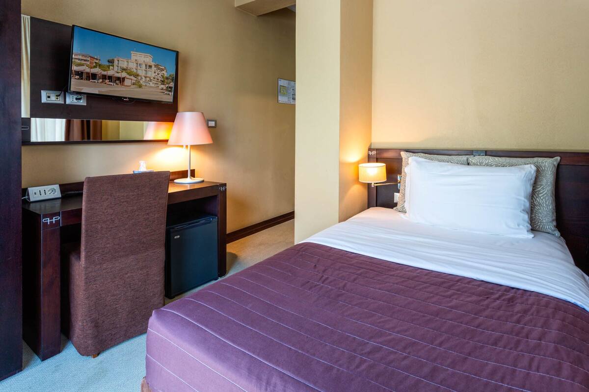 A hotel room with a bed, a desk, a chair, a TV, and a lamp.