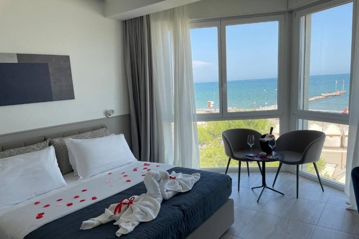 A hotel room with a bed, a balcony with a view of the sea, and a table with a glass of wine.