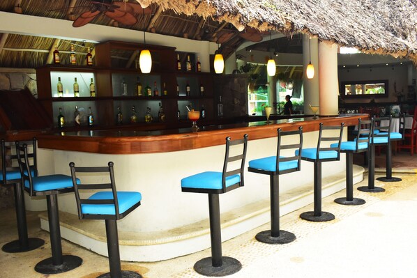 Zona de bar con techo de paja, taburetes y estantes llenos de botellas.