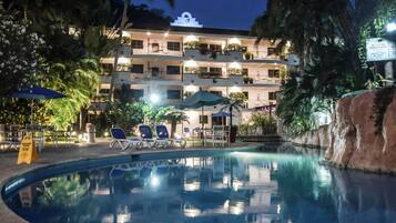 Una piscina con sillas de descanso, palmeras y un edificio con balcones al fondo.