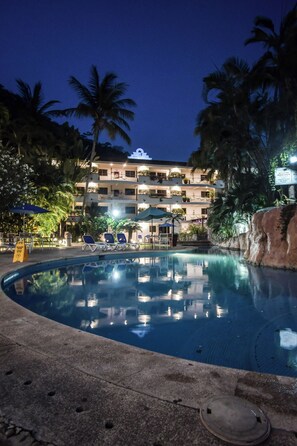 Ein Swimmingpool mit Liegestühlen, Palmen und ein Gebäude mit Balkonen im Hintergrund.