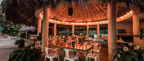 An outdoor dining area with a thatched roof, white chairs, and tables.