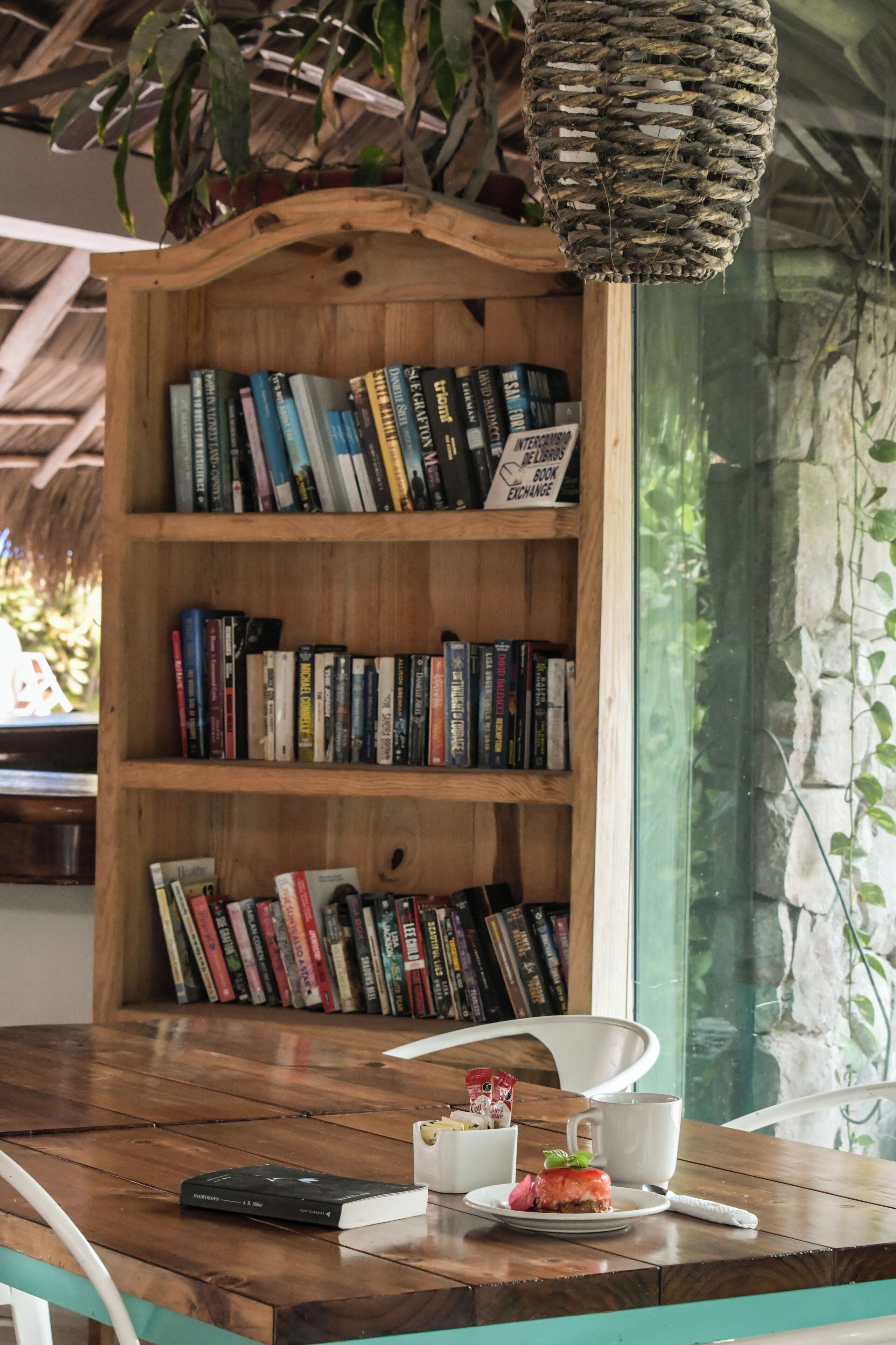 Una estantería de madera llena de libros, una mesa con un plato de comida y un mando a distancia, y una lámpara colgante.
