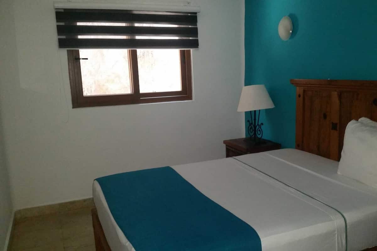A bedroom with a bed, a window with blinds, a nightstand with a lamp, and a teal accent wall.