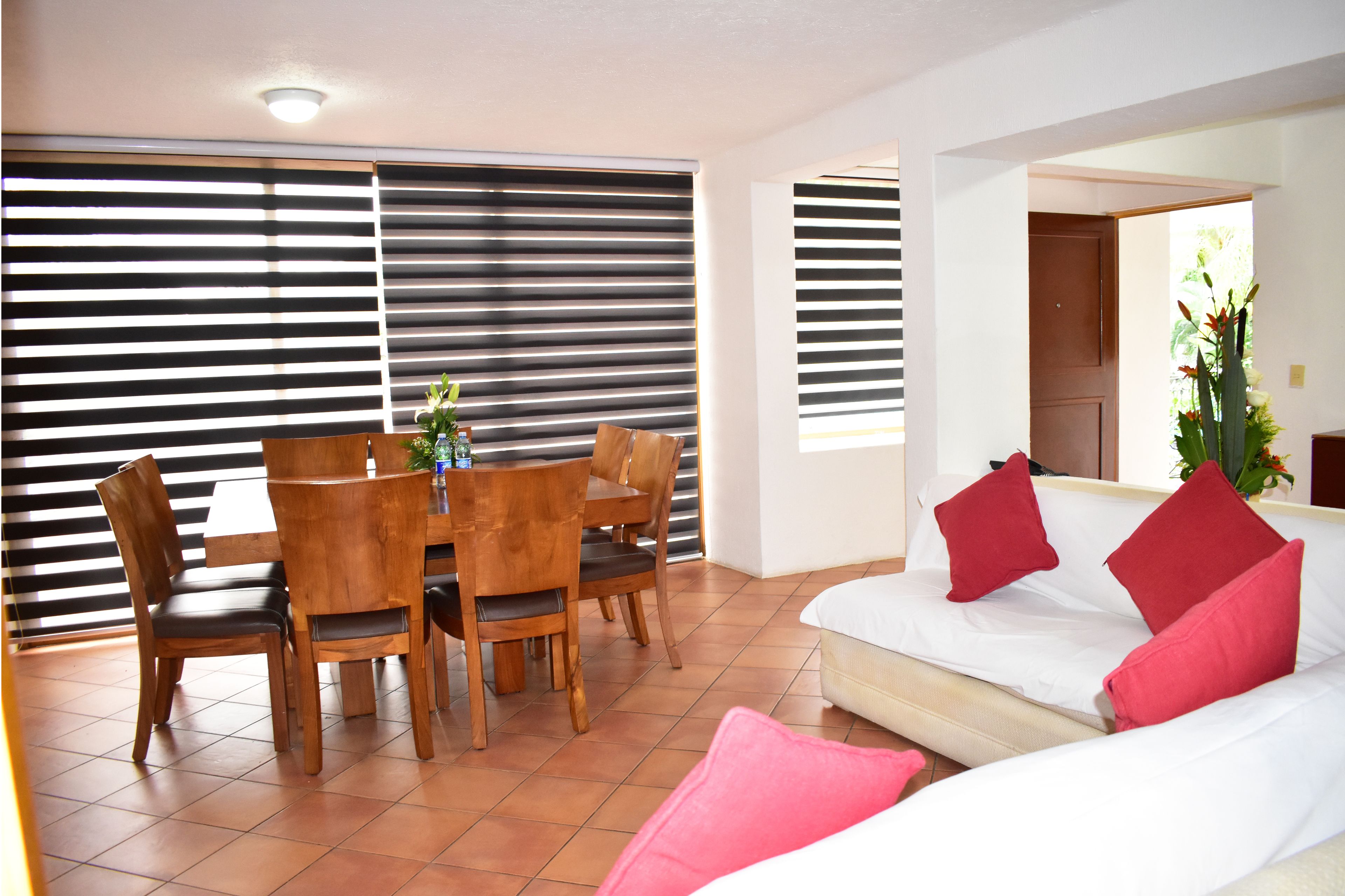 A living room with a dining table and chairs, a sofa with red cushions, and a large window with blinds.