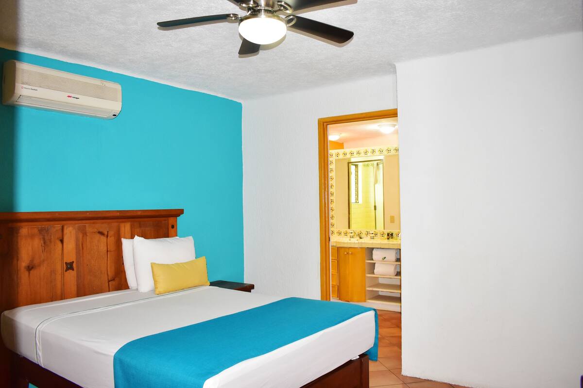 A hotel room with a bed, a ceiling fan, an air conditioner, and a bathroom visible through an open door.
