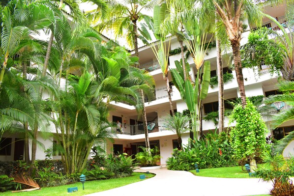 A multi-story hotel with lush greenery, including palm trees and various plants. There are balconies on each floor, and a pathway leading to the entrance.