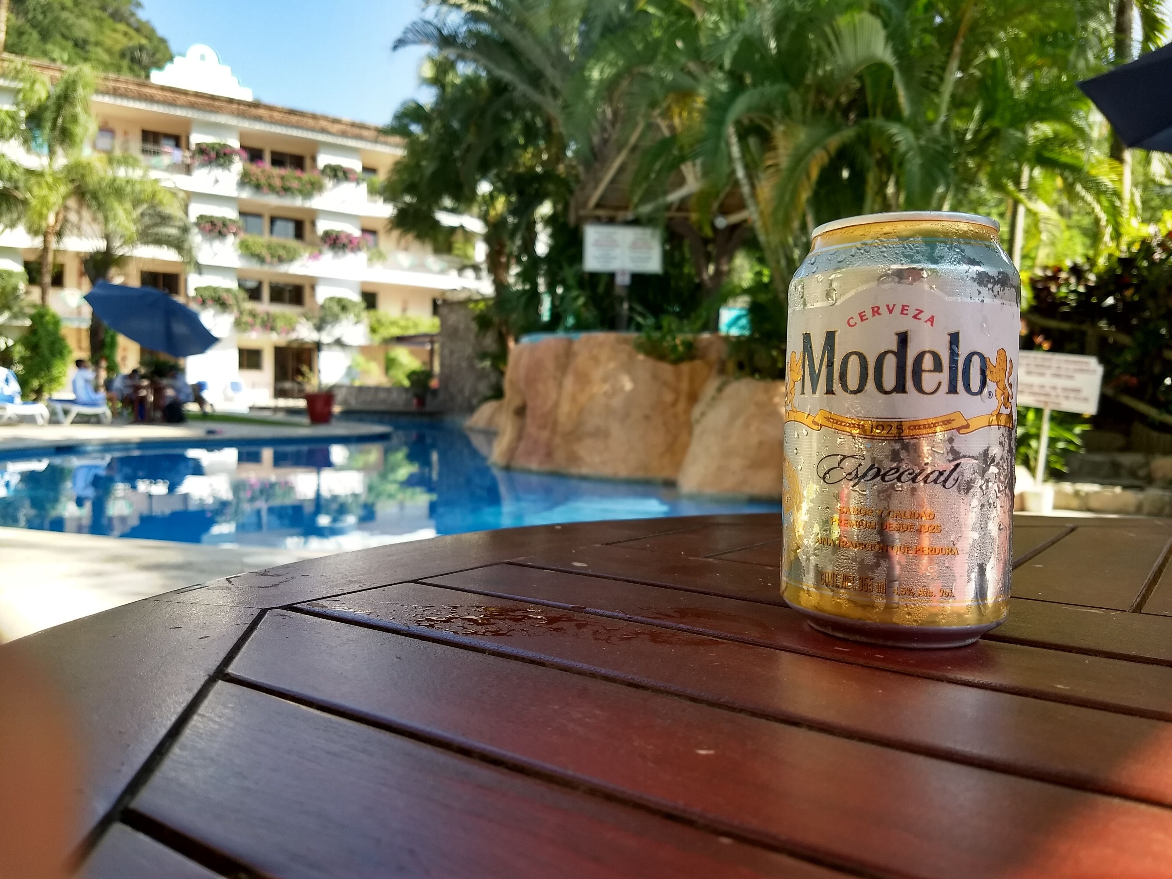 Une canette de bière Modelo sur une table près d’une piscine, avec un bâtiment en arrière-plan.