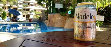 A can of Modelo beer on a table near a pool with a building in the background.