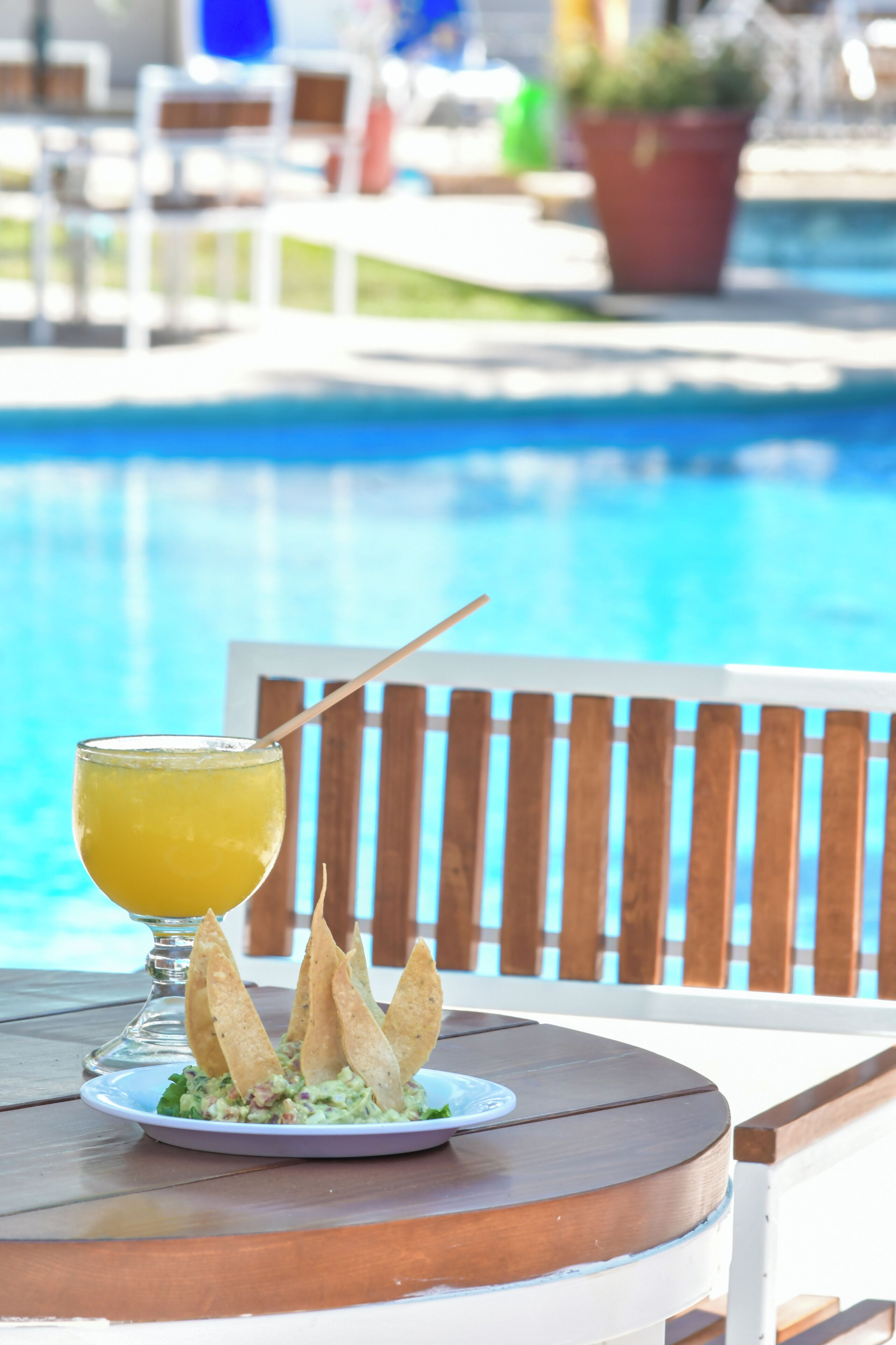 A poolside table with a drink and chips, a wooden chair, and a pool in the background.