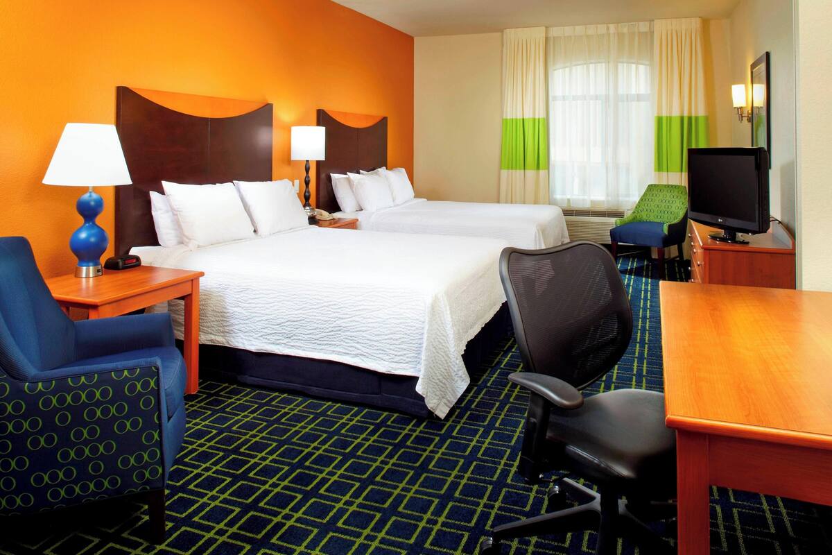 A hotel room with two beds, a desk, a chair, a TV, and a lamp.