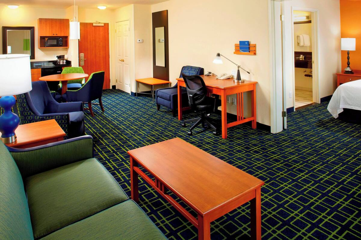A hotel room with a sofa, a desk, a chair, a small table, a lamp, a microwave, a mirror, a closet, and a bed.
