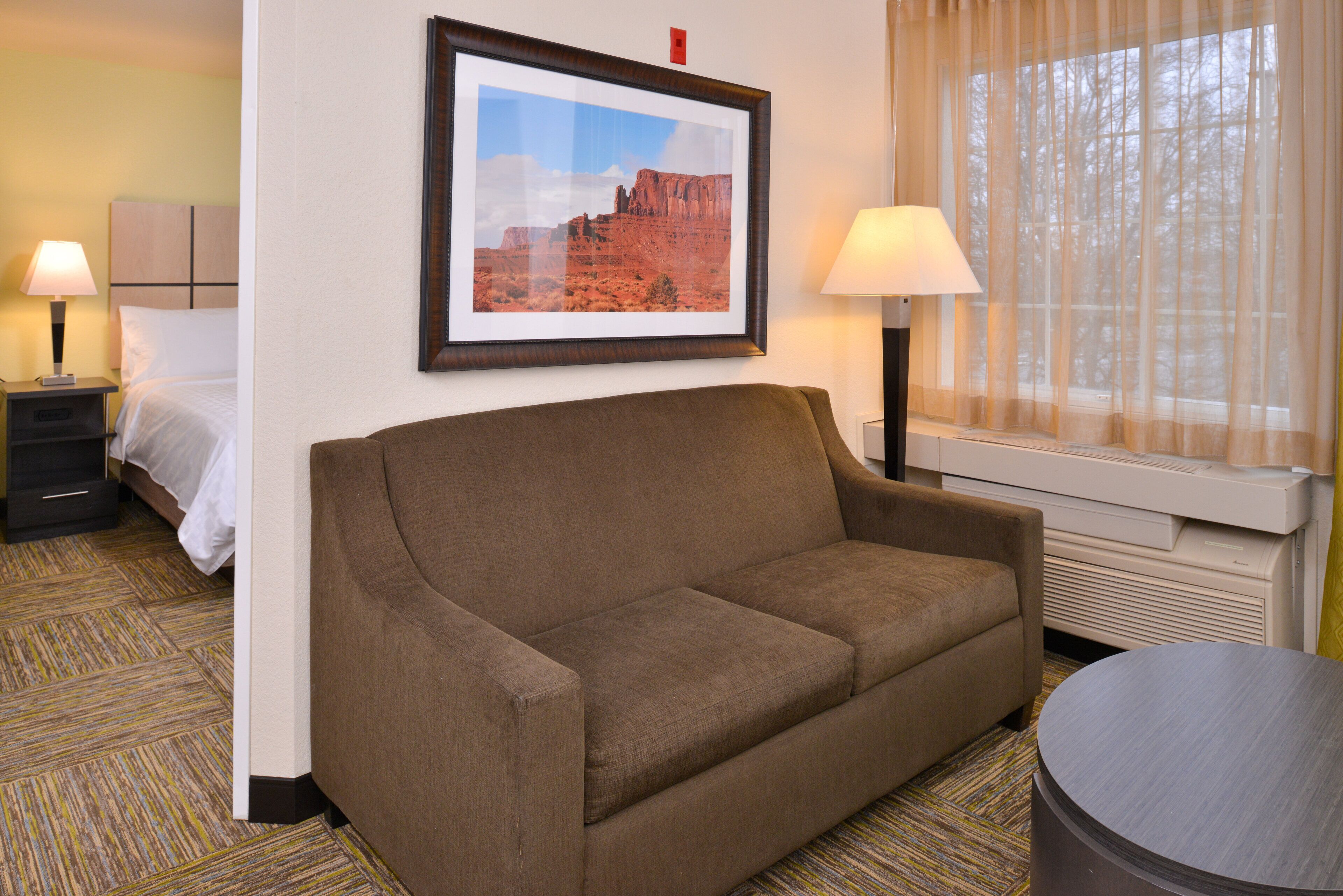 Ein Hotelzimmer mit braunem Sofa, einem Bett mit weißer Bettwäsche, zwei Nachttischlampen, einem gerahmten Bild an der Wand und einem Fenster mit transparenten Vorhängen.