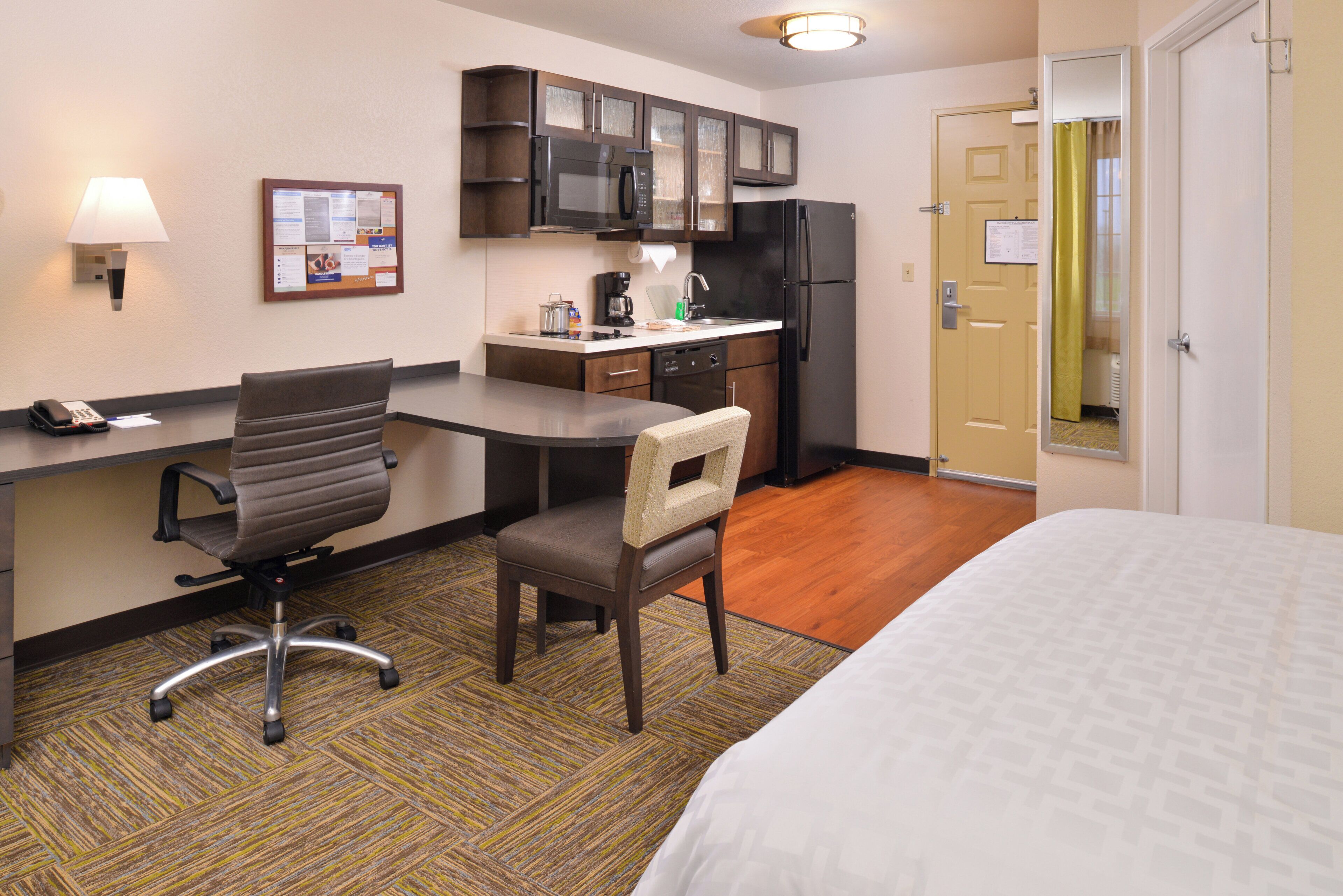 A hotel room with a bed, a desk with a chair, a kitchenette, and a door.