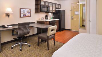 A hotel room with a bed, a desk with a chair, a kitchenette, and a door.