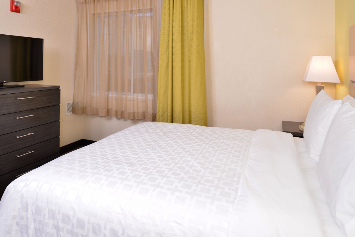 A hotel room with a bed, a TV on a dresser, a lamp, and a window with sheer curtains.