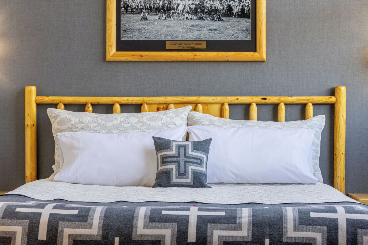 A bed with a wooden headboard, white and patterned pillows, and a cross-shaped pillow. A framed photo is on the wall above the bed.