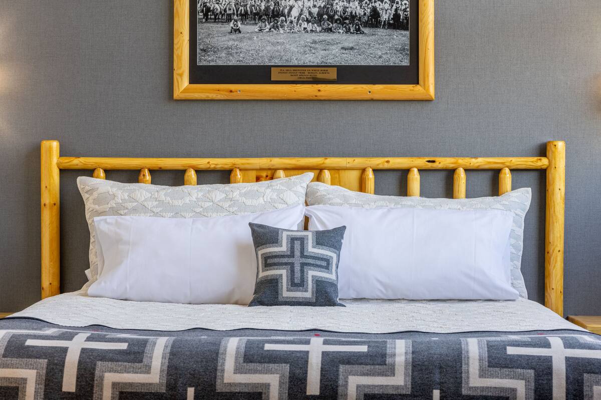 A bed with a wooden headboard, white and patterned pillows, and a cross-shaped pillow. A framed photo is on the wall above the bed.