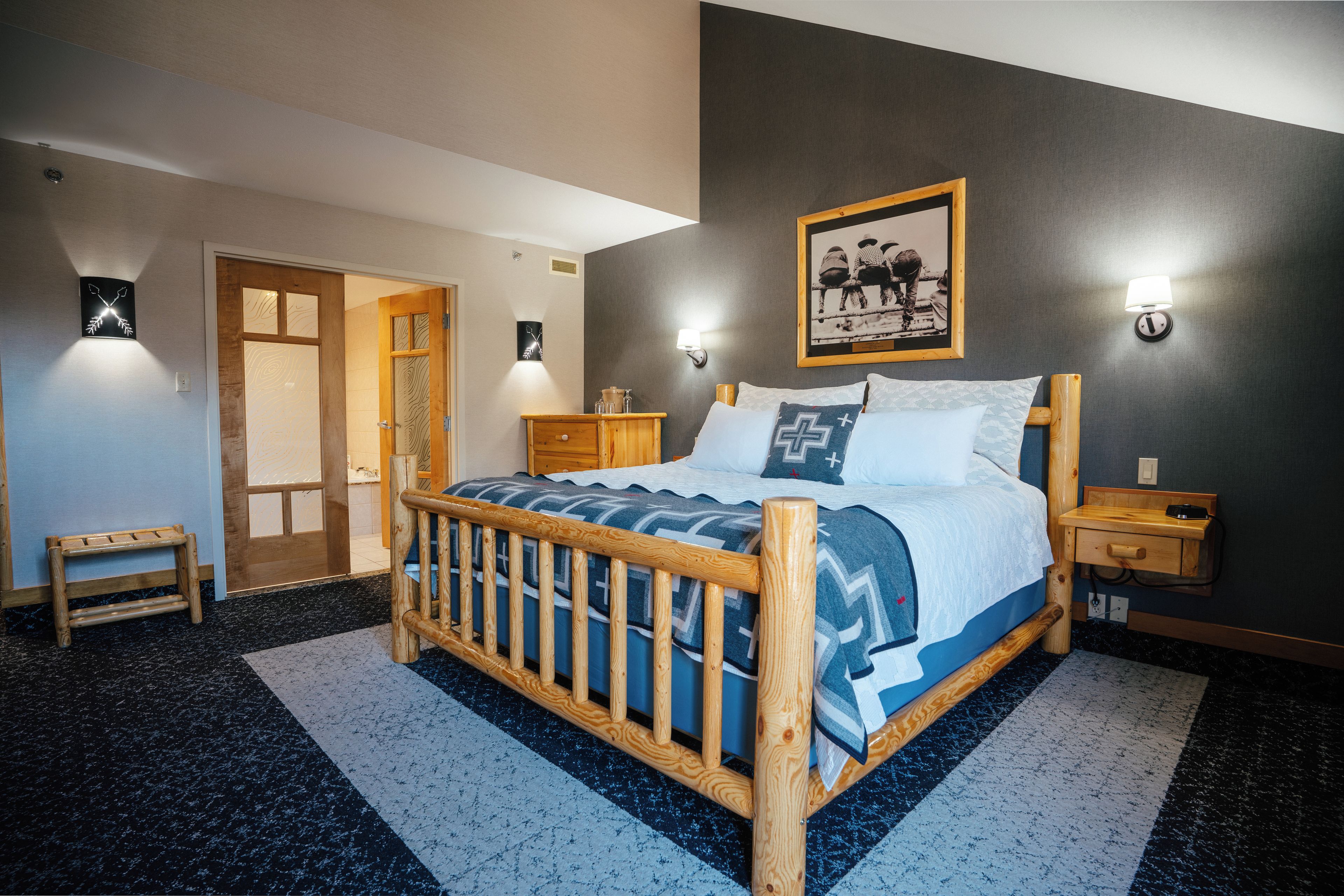 A bedroom with a wooden bed, a nightstand, a bedside table, a wall-mounted lamp, and a framed picture.