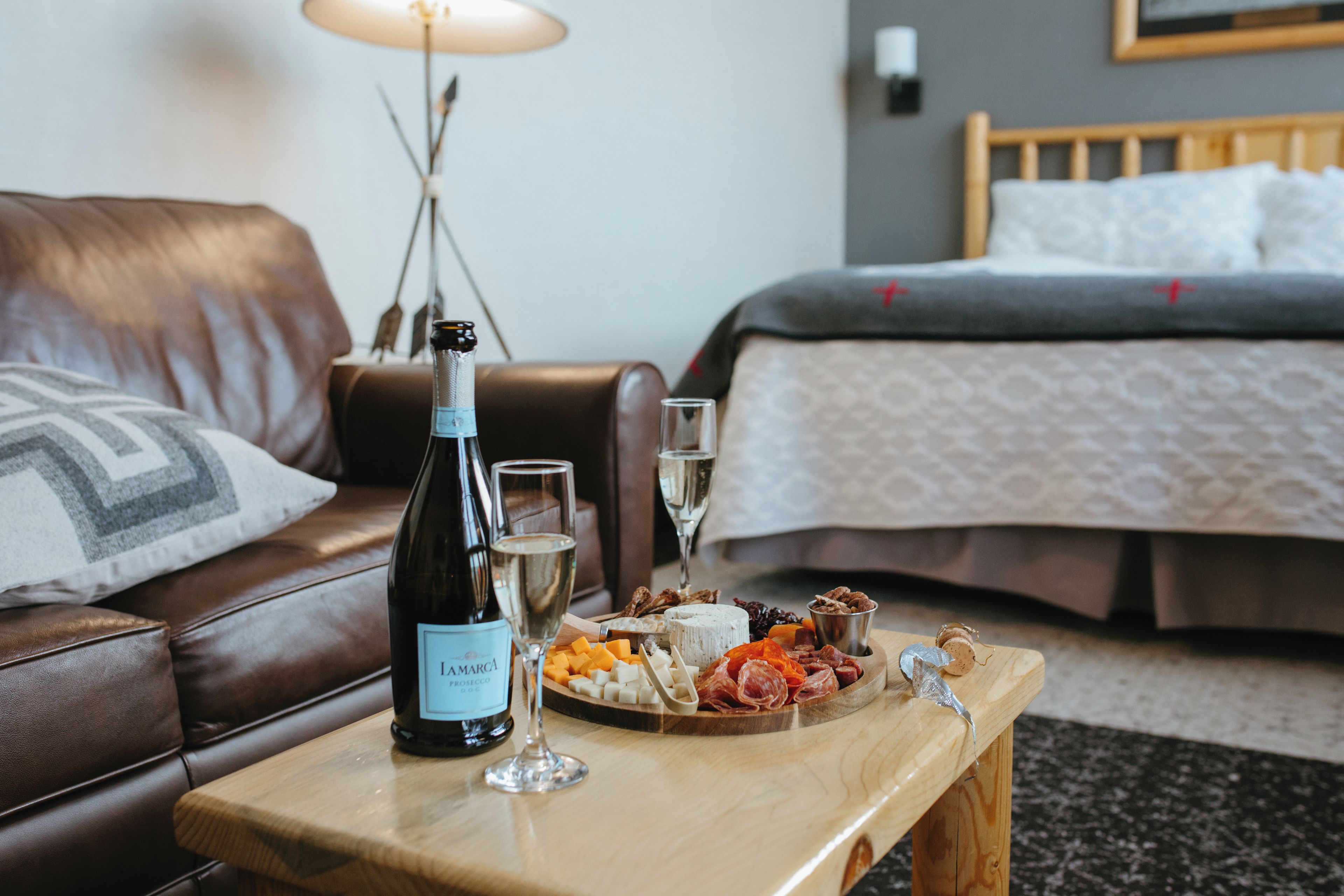 Habitación de hotel con una mesa de centro de madera que sostiene una botella de vino, copas y una tabla de quesos.
