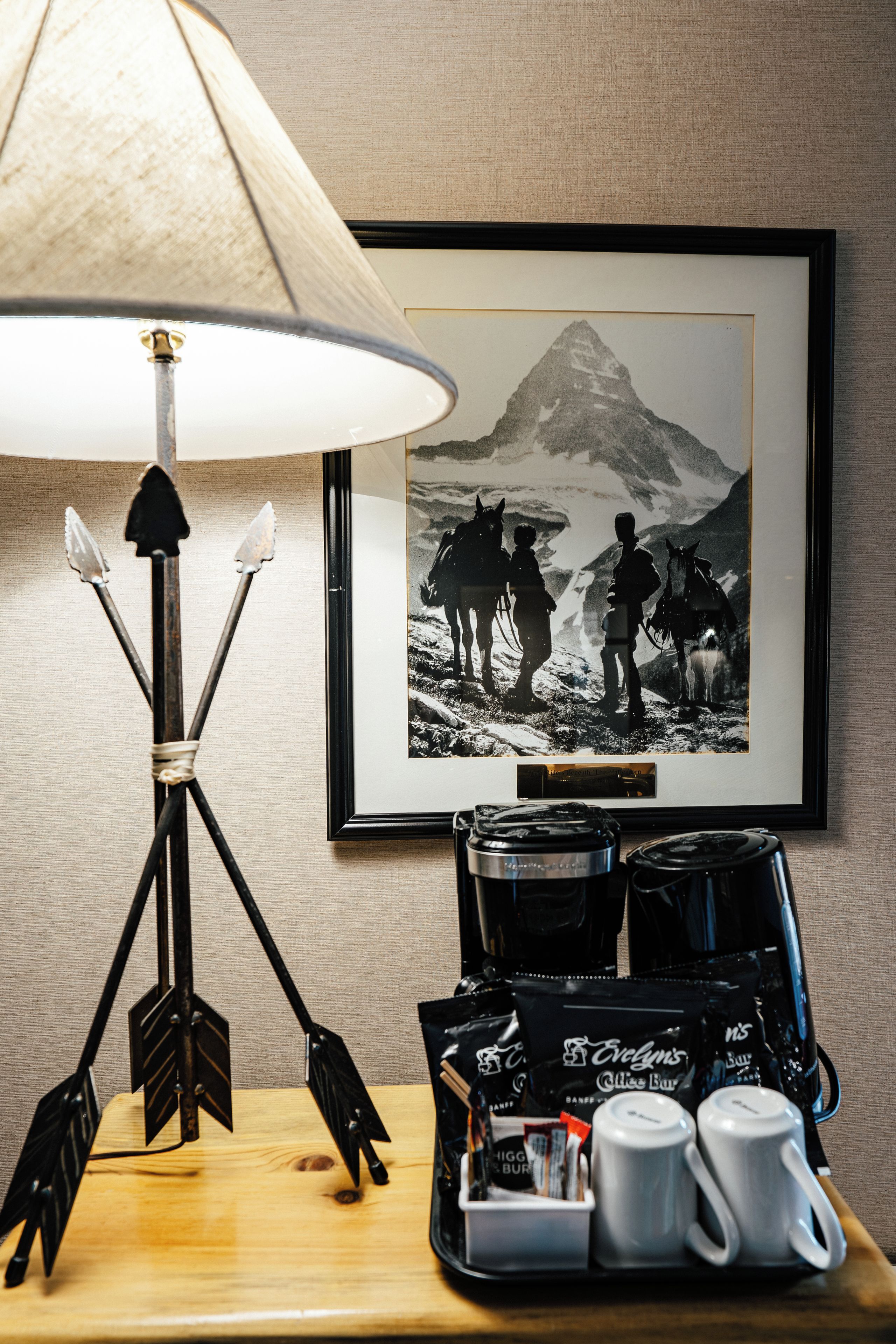 A table with a coffee maker, cups, and bags of coffee, a lamp, and a framed picture of a mountain scene.