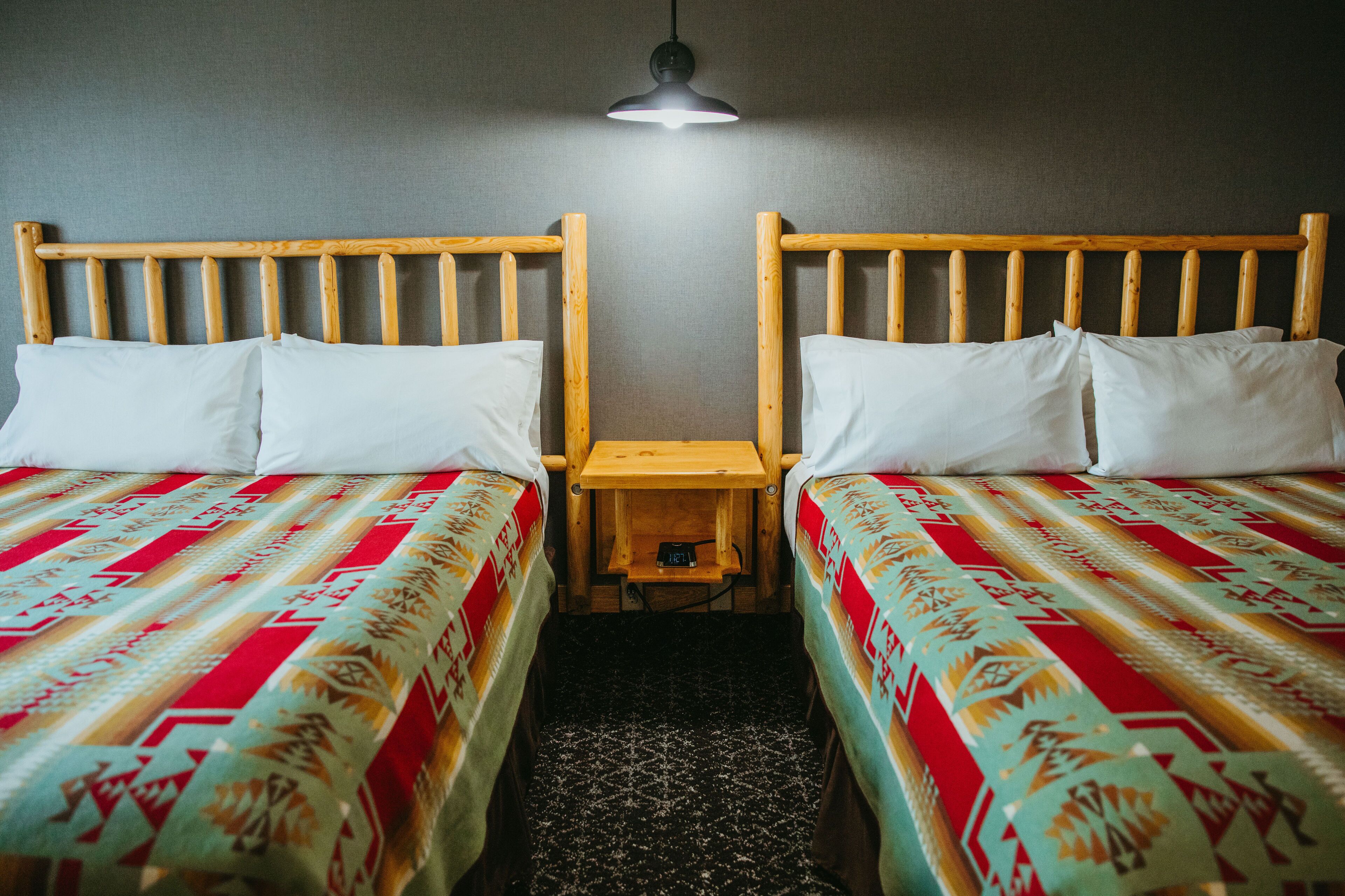 A room with two wooden beds, each with a red and white patterned bedspread, a small wooden nightstand, and a single overhead light.