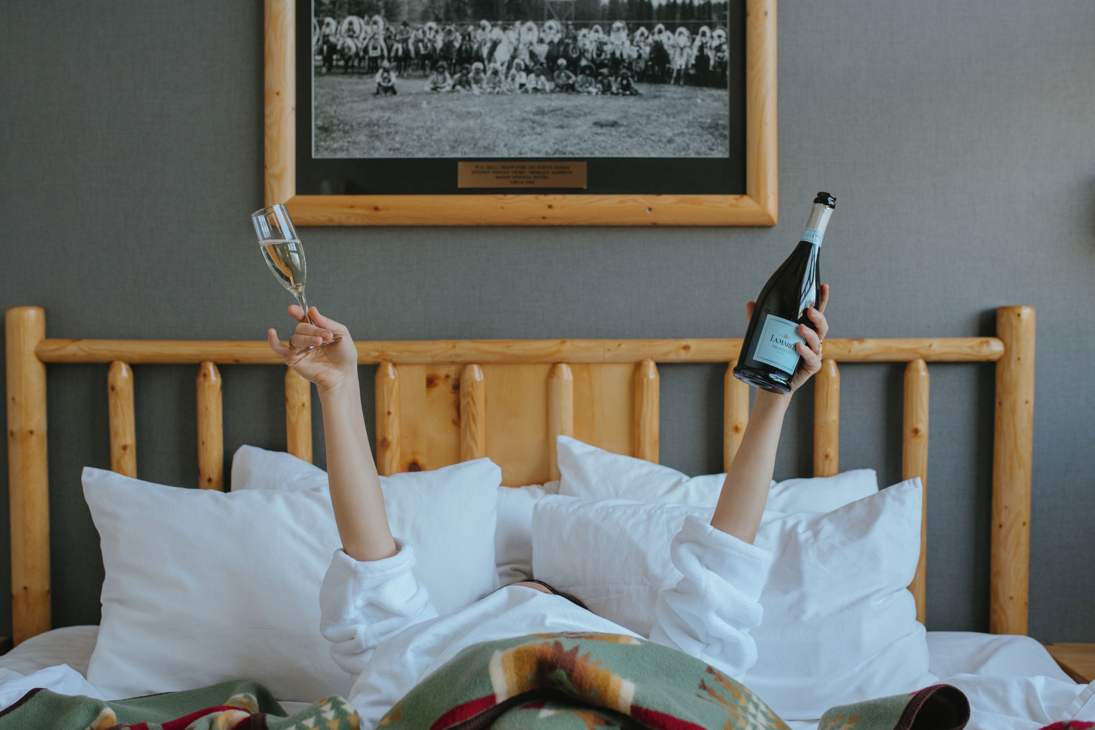 Una persona sdraiata su un letto con una bottiglia e un bicchiere di vino, una foto incorniciata al muro e una testiera in legno.