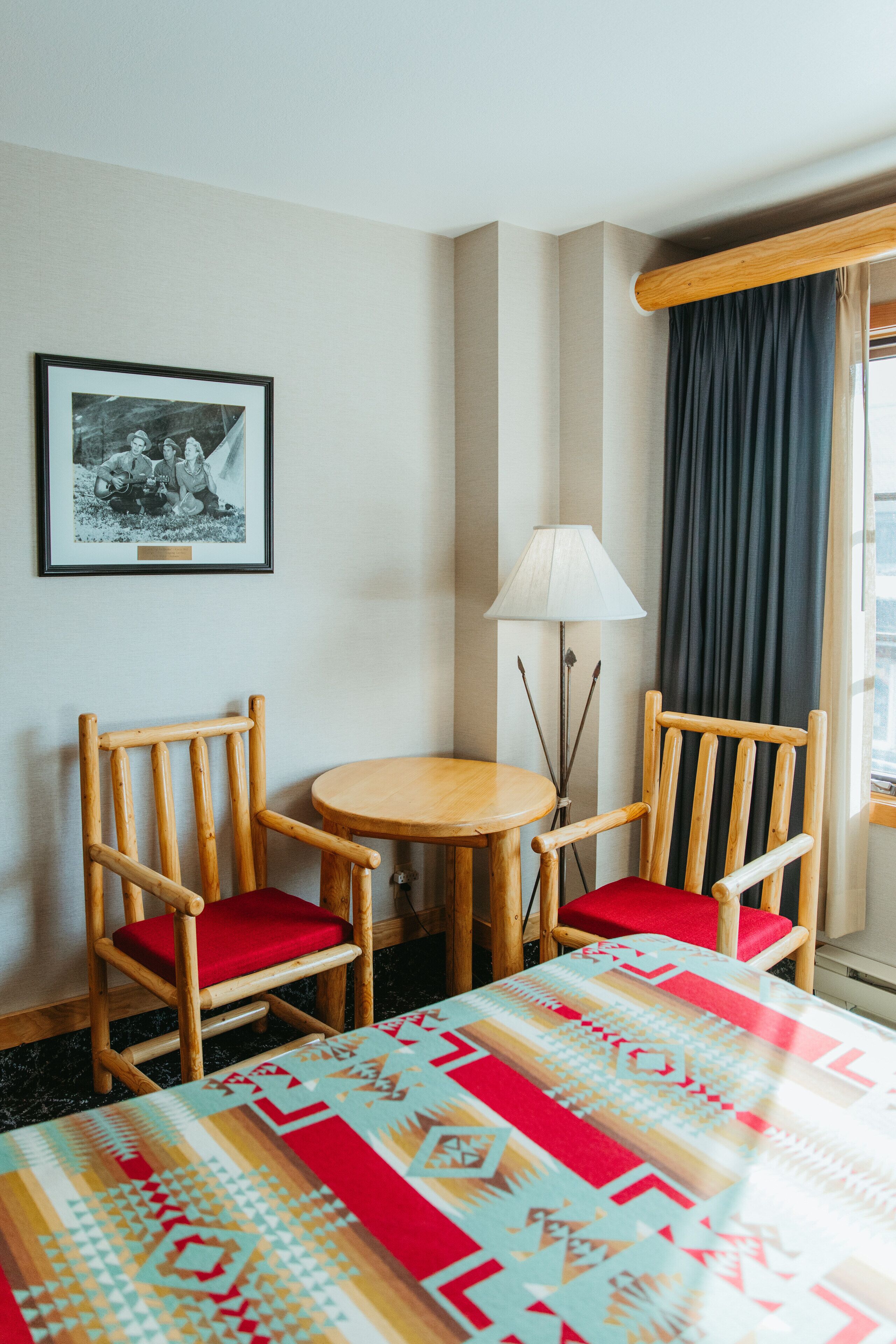 A hotel room with a bed, two wooden chairs with red cushions, a small round table, a lamp, and a framed picture on the wall.