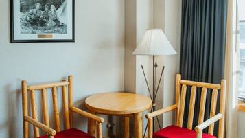 A hotel room with a bed, two wooden chairs with red cushions, a small round table, a lamp, and a framed picture on the wall.