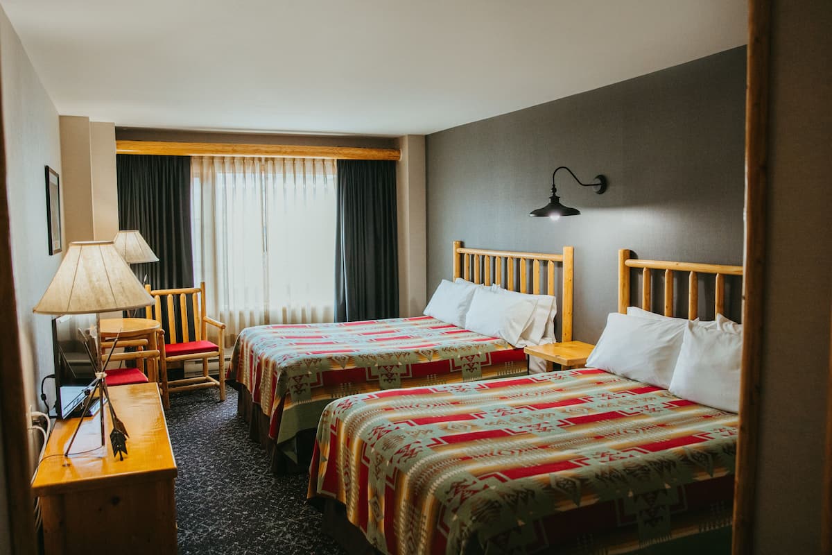 A hotel room with two beds, a desk with a lamp, a chair, and a window with curtains.