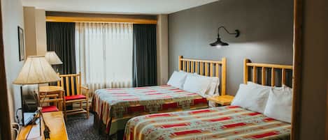 Habitación de hotel con dos camas, un escritorio con lámpara, una silla y un ventanal con cortinas.