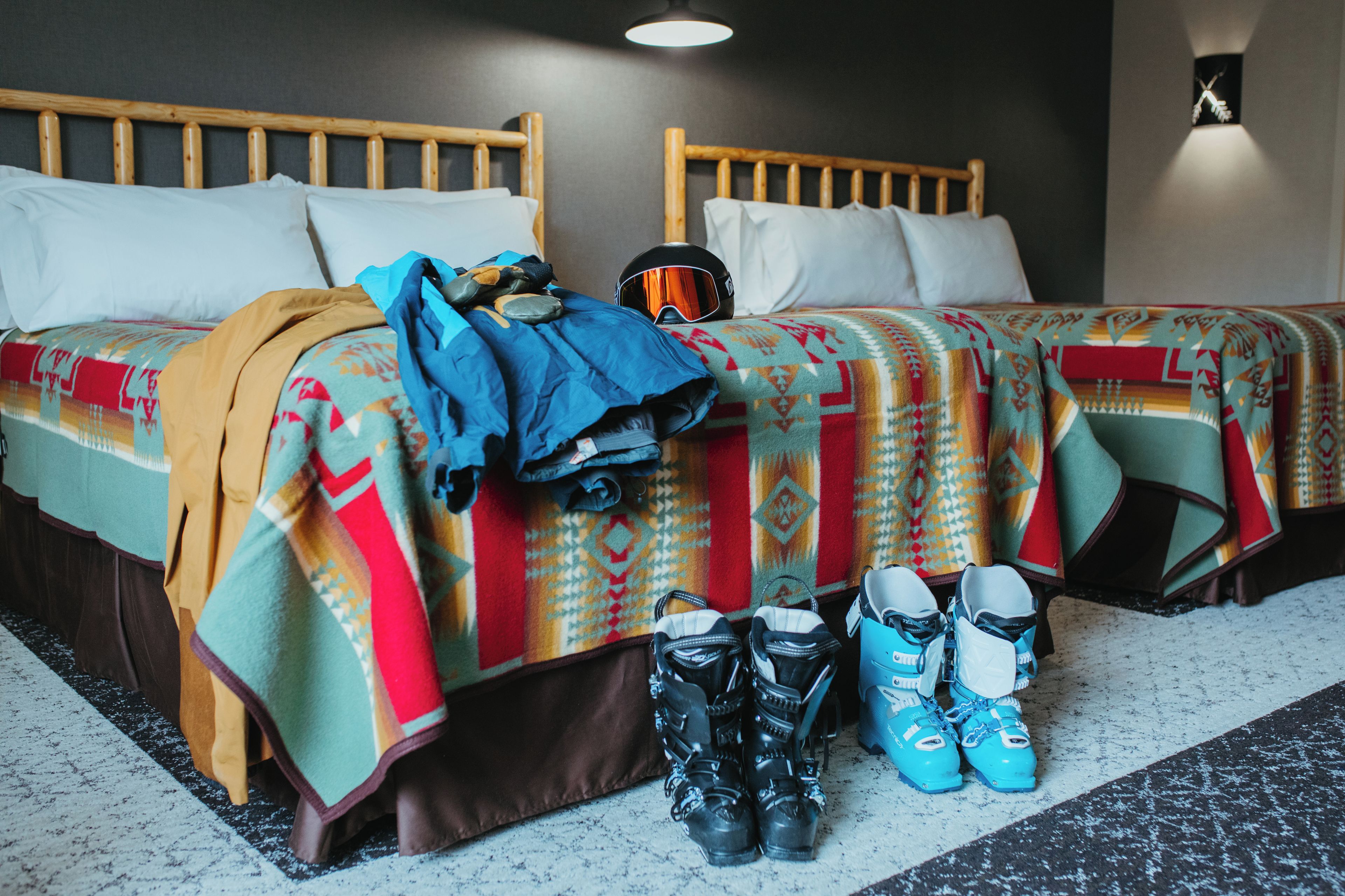 Une chambre d’hôtel avec deux lits, une tête de lit en bois, une lampe fixée au mur, et une paire de patins à roulettes et de chaussures posées sur le sol.