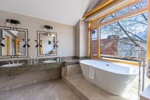 A modern bathroom with two sinks, a freestanding bathtub, and a large window offering a view of trees and buildings.