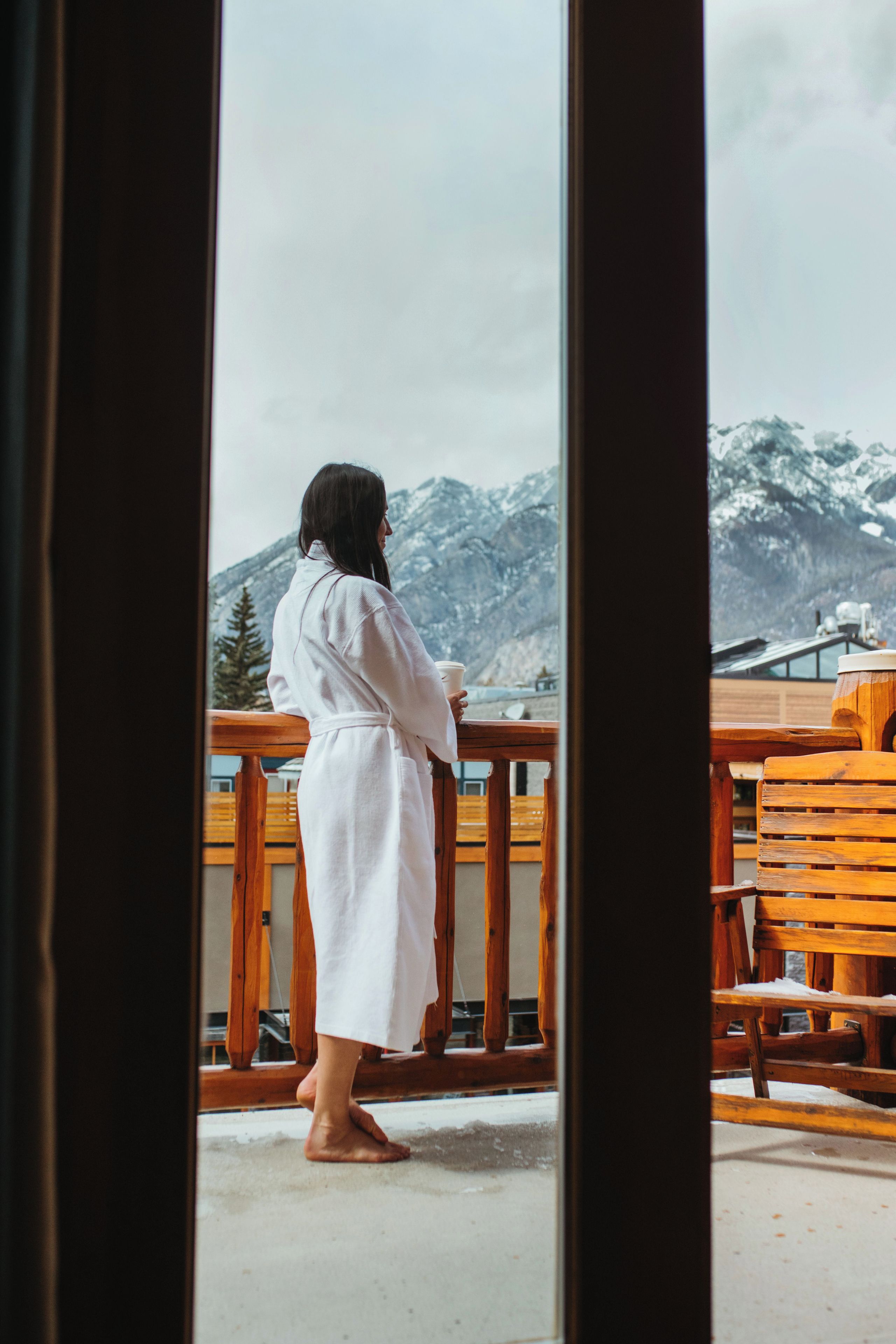 A person in a white robe standing on a balcony with wooden railings, overlooking a mountainous landscape.