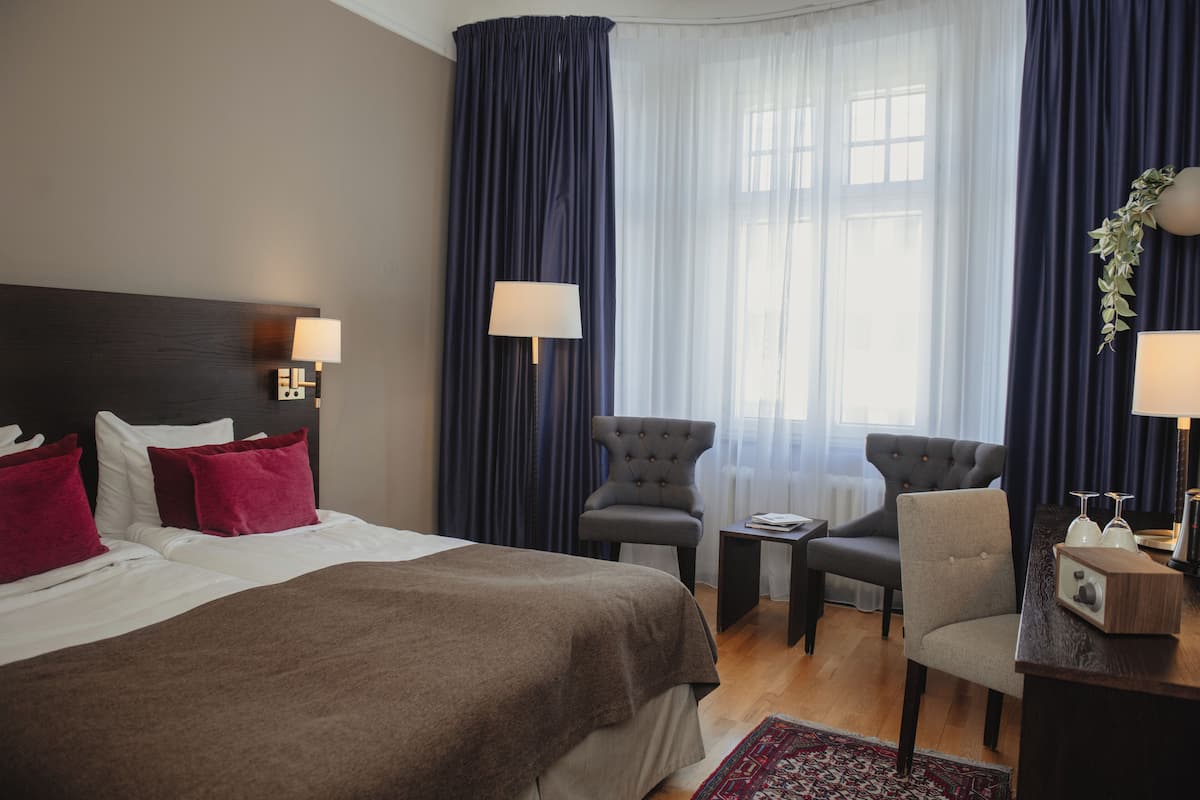 Quarto de hotel com uma cama grande, duas poltronas, uma mesa pequena e uma escrivaninha com abajur.