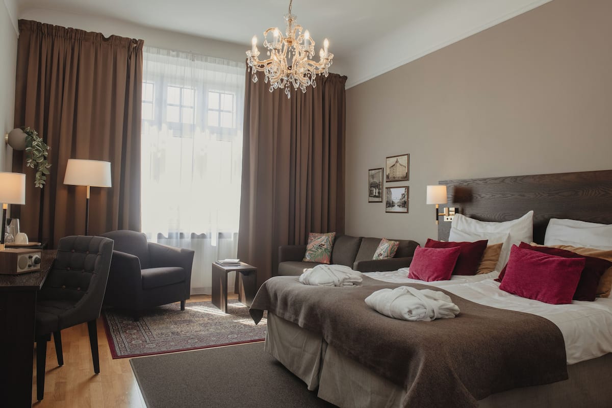 Quarto de hotel com uma cama grande, um sofá, uma escrivaninha com abajur e um candelabro.