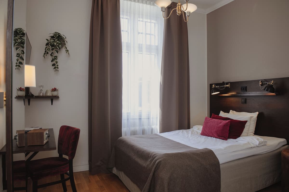 Quarto de hotel com cama, escrivaninha, cadeira, mesa de cabeceira, abajur e janela com cortinas.