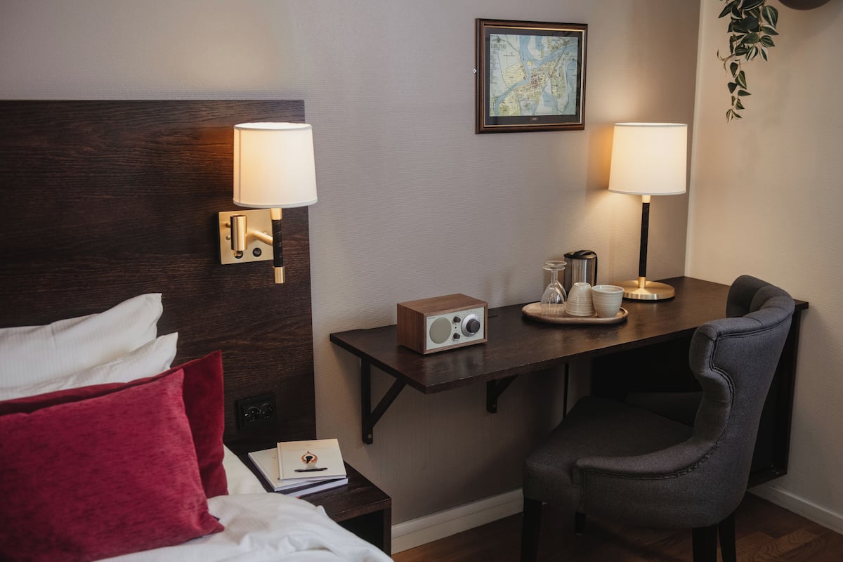 Quarto de hotel com cama, mesa de cabeceira, escrivaninha, cadeira, luminárias e um mapa emoldurado na parede.