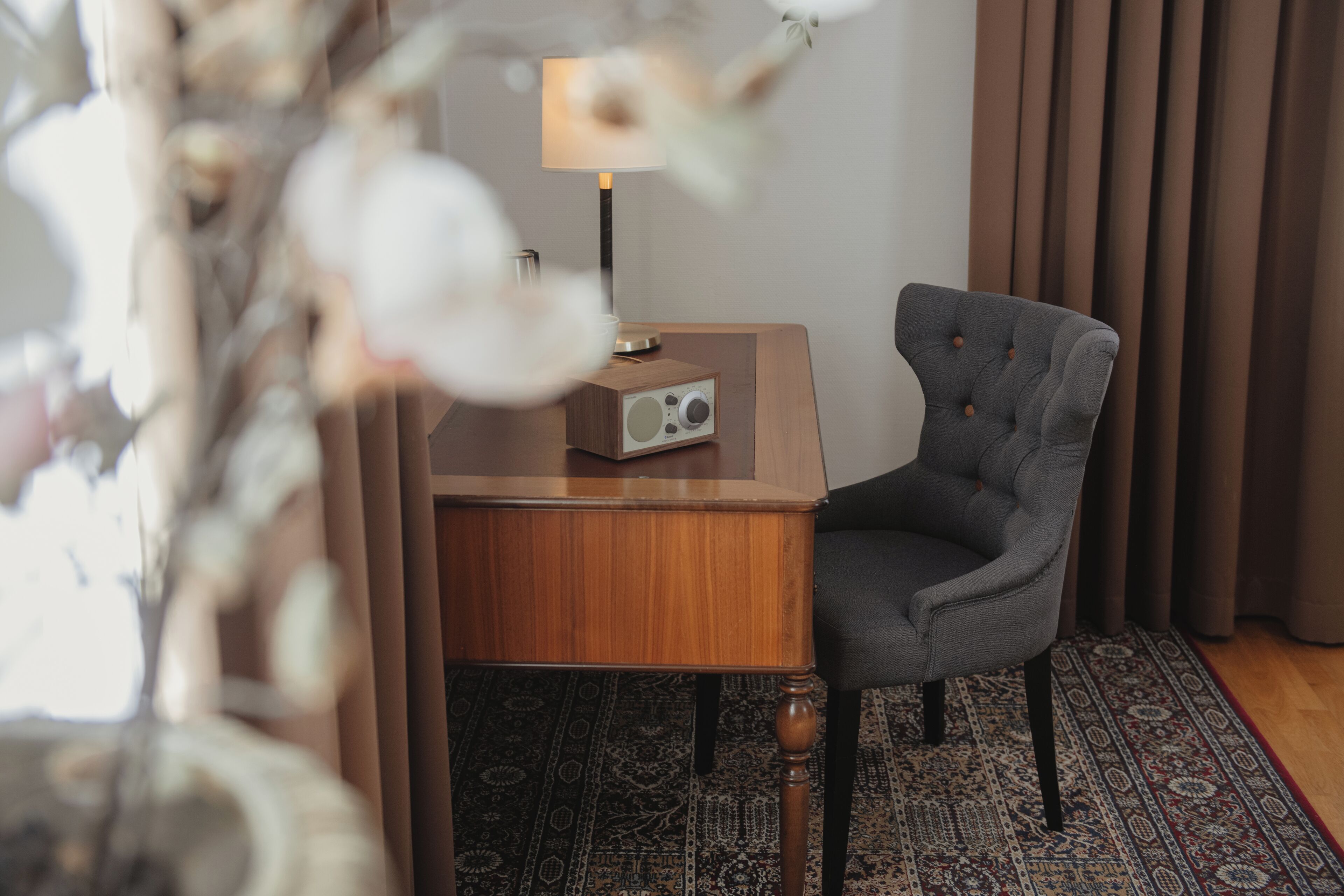 A room with a wooden side table, a grey upholstered chair, a radio, a lamp, and a patterned rug.