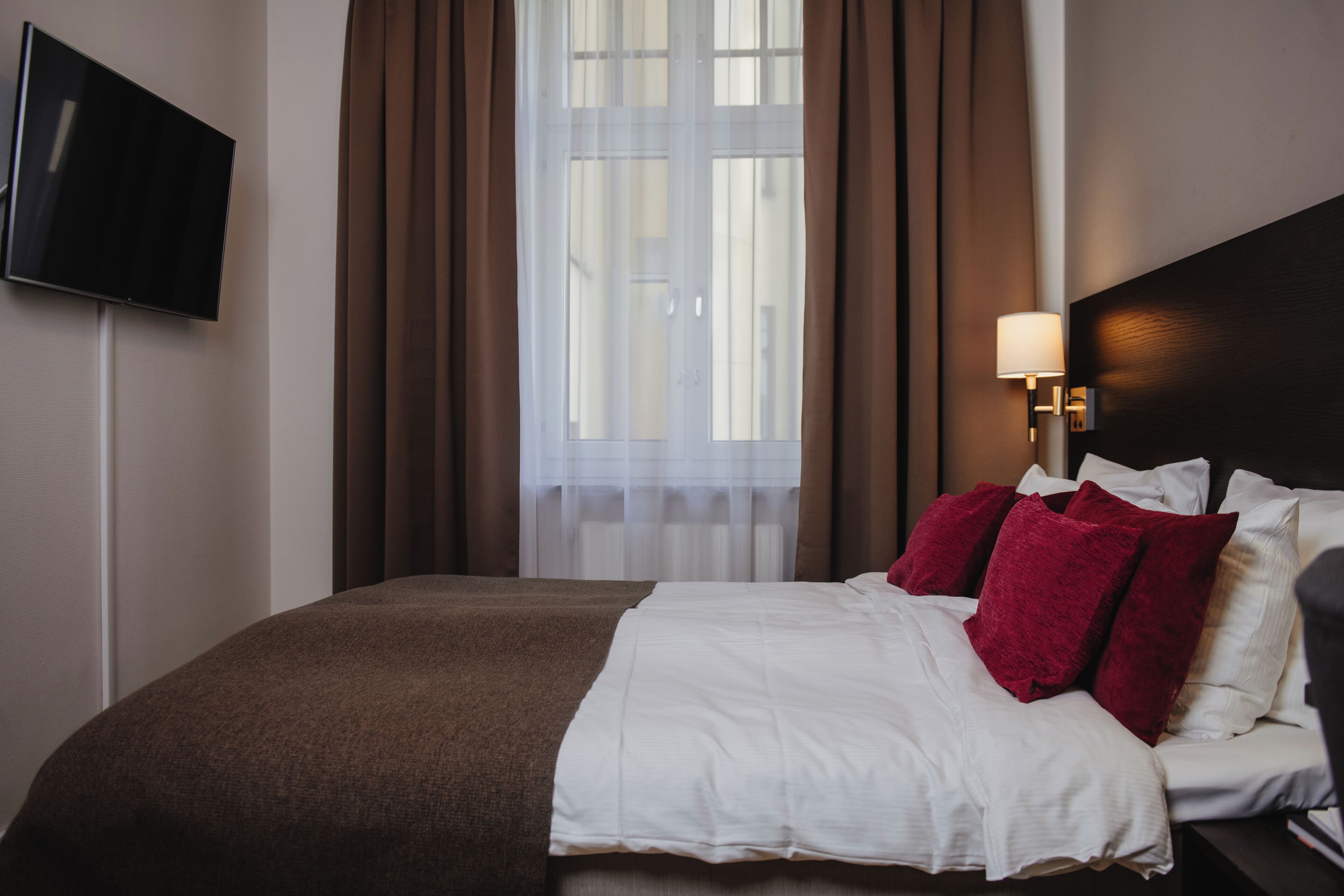 A neatly made bed with white and red pillows, a brown blanket, and a TV mounted on the wall.