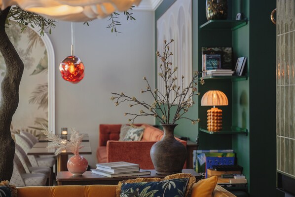 A cozy living room with a sofa, a coffee table with books, a vase with dried branches, a red ornament, and a green bookshelf.