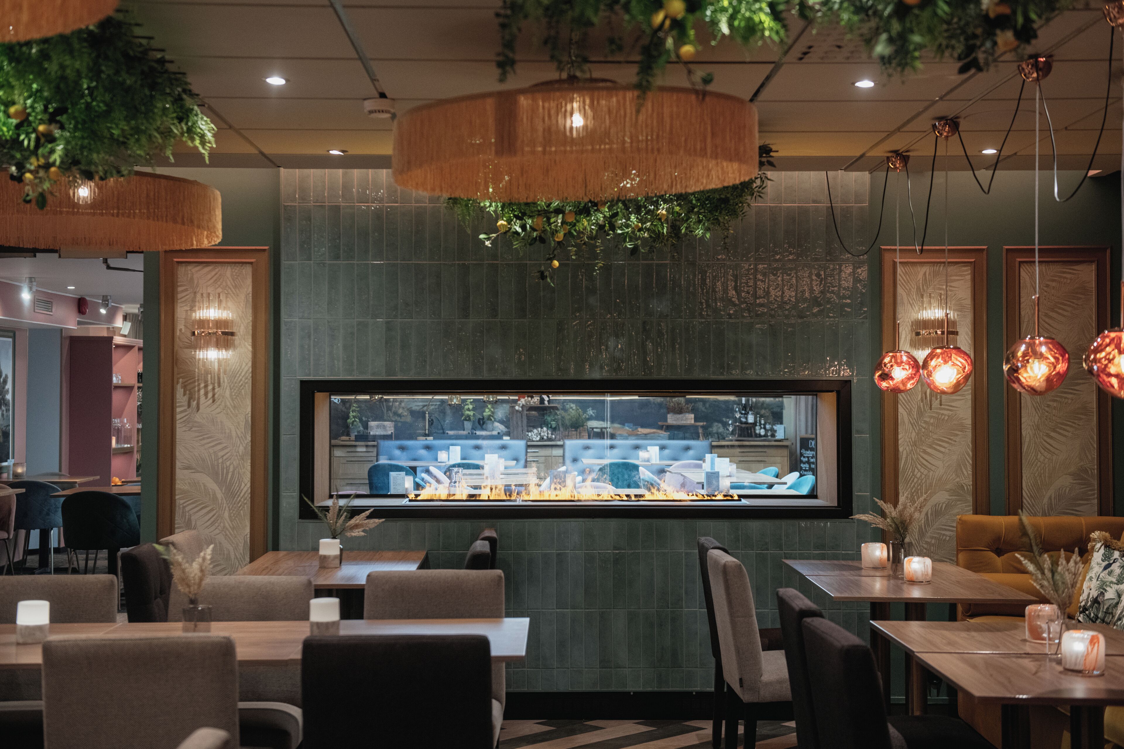 A restaurant interior with wooden tables, cushioned seating, hanging plants, and a large window overlooking the street.