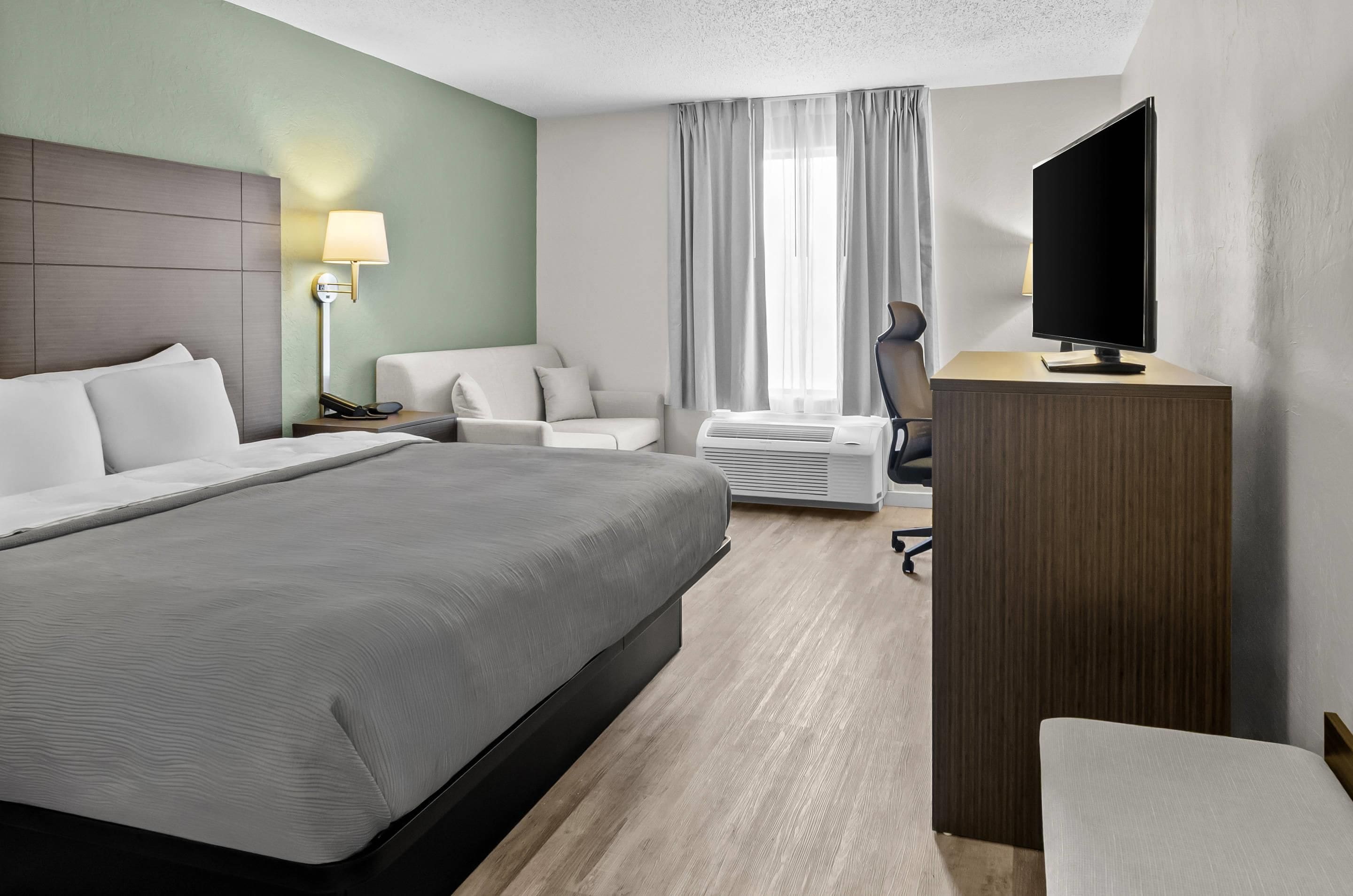 Habitación de hotel con una cama grande, un escritorio con televisor de pantalla plana, una silla, una lámpara de escritorio y una ventana con cortinas.