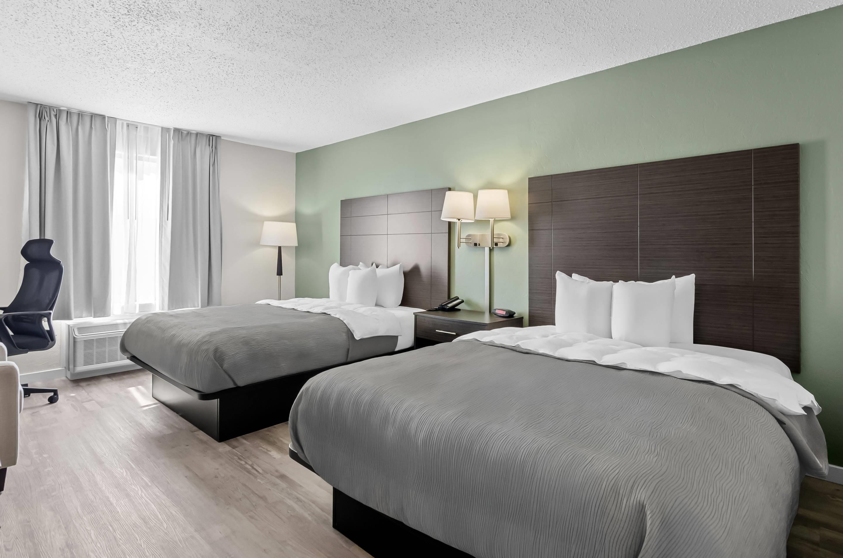A hotel room with two beds, a desk, a chair, and a lamp.