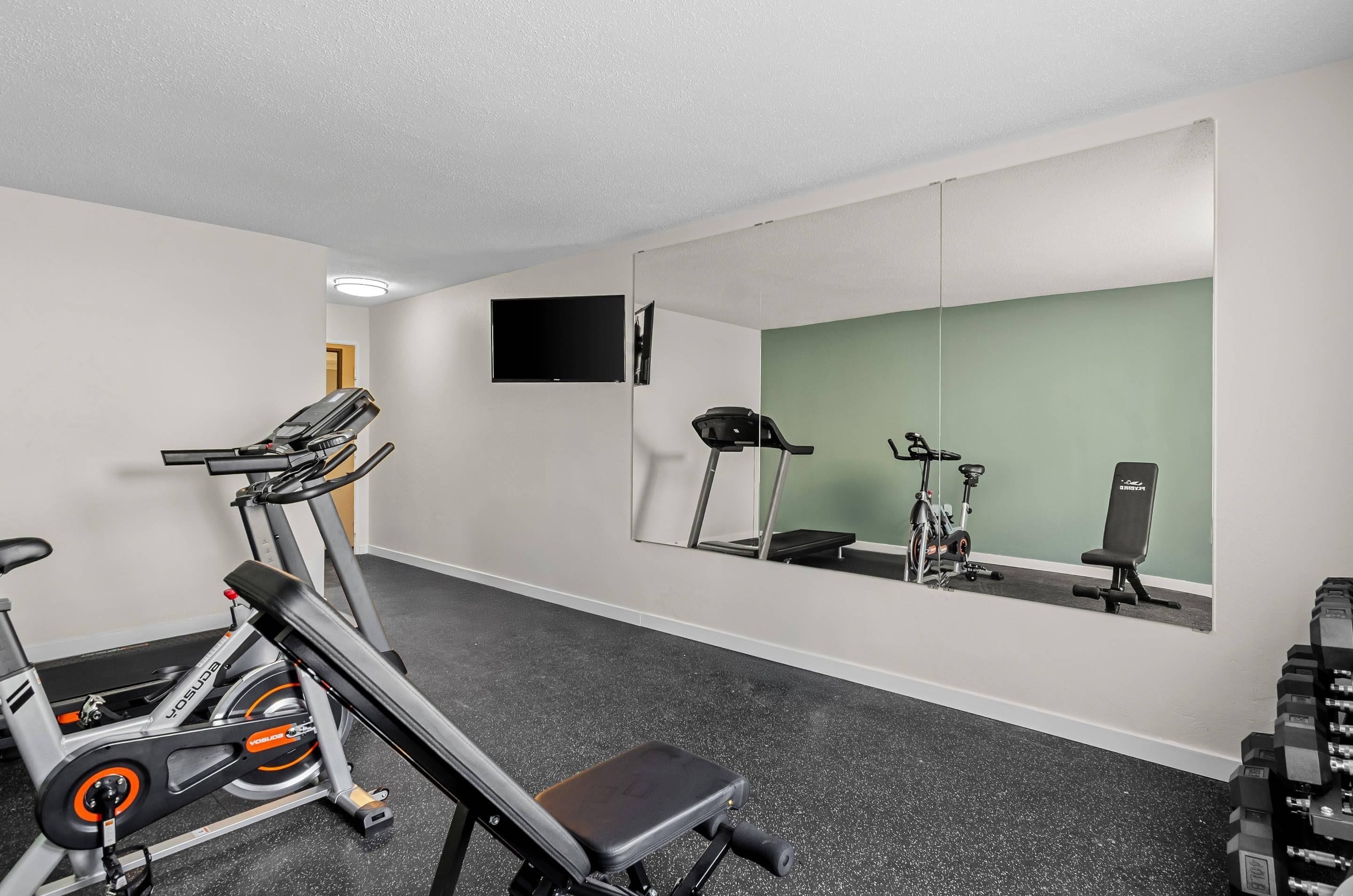 Uma academia com equipamentos de cardio, um banco e um espelho refletindo o ambiente.