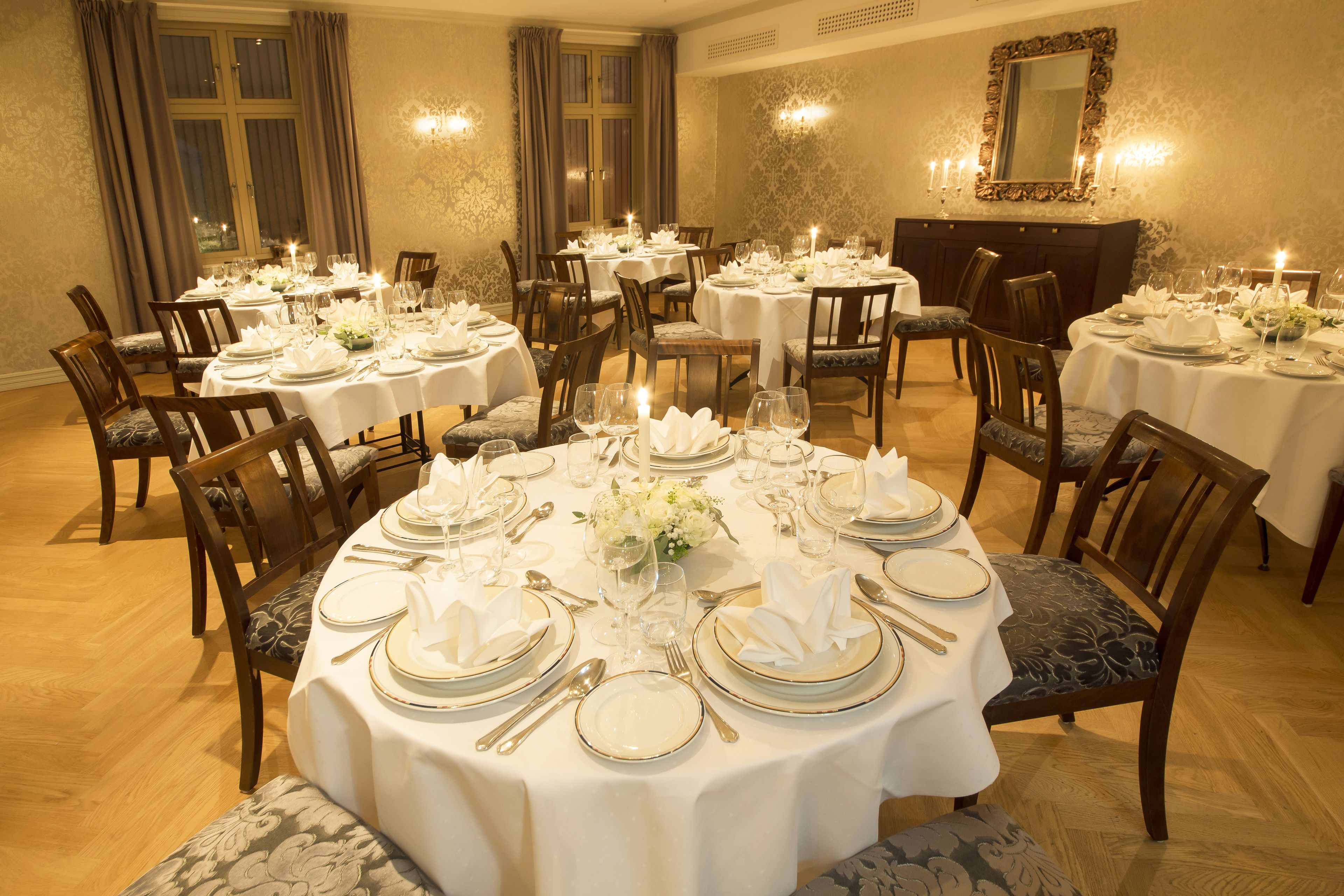 A dining area with multiple round tables set for a formal meal, featuring white tablecloths, silverware, and floral centerpieces.