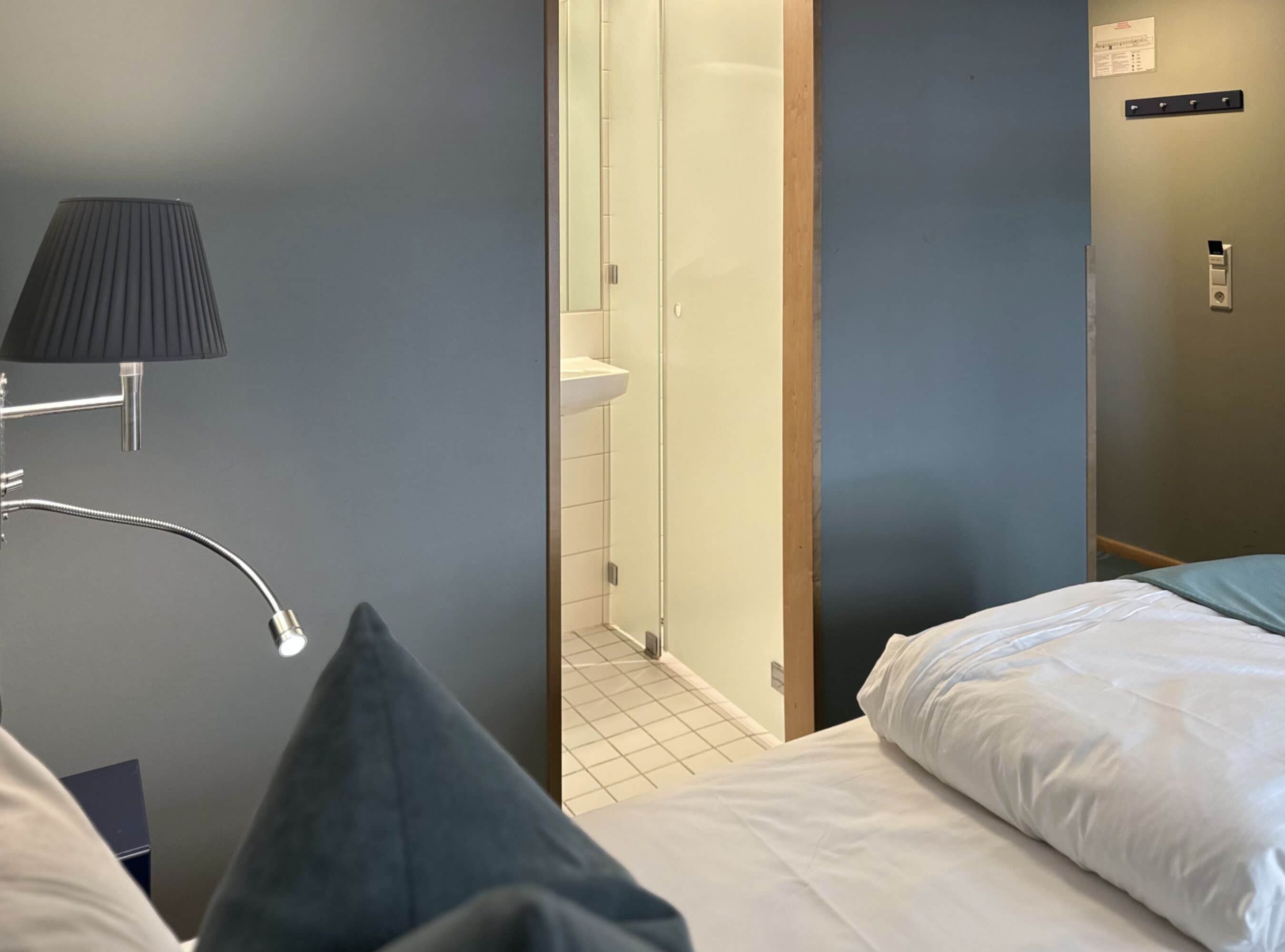 Habitación de hotel con cama, mesita de noche, lámpara, baño con lavamanos y espejo.